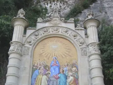 Monumental Rosary of Montserrat with a colorful Pentecost mosaic in a stone arch, seen behind a black metal fence.