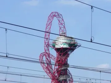 Orbit Tower with a red twisting lattice structure and circular platform near the top, seen behind overhead power lines and railway tracks.