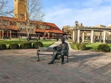 Seated Lincoln sculpture on a bench in a brick-paved public square with landscaped shrubs and buildings nearby.