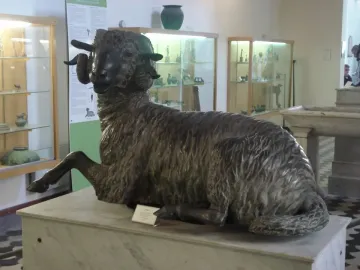 Bronze ram sculpture with curled horns resting on a stone pedestal in a museum gallery.