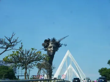 Reminiscencia, a dark metallic bust of a woman with a sculpted floral crown, stands outdoors against a blue sky.