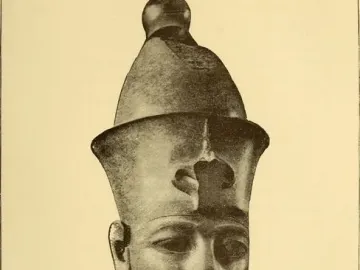 Colossal head of Amenhotep III displayed on a white pedestal in a museum gallery, with a smaller stone fragment beside it.