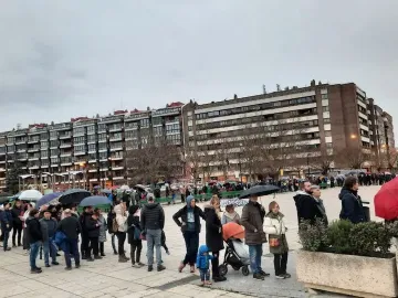 A large crowd gathers on a paved plaza under a cloudy sky, with umbrellas open and apartment buildings in the background.