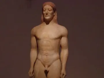 Kroisos Kouros, a nude male Greek statue with braided hair standing on a base in a museum setting.
