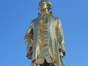 Golden statue of Alexander Hamilton in historical attire on a dark pedestal against a blue sky.