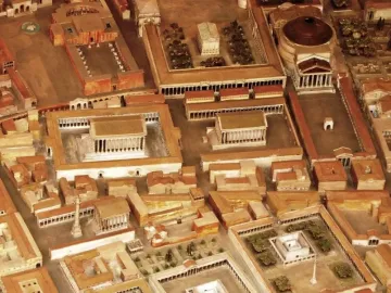 An aerial view of a Roman city scale model with a prominent white column in an open plaza, representing the Antoninus Pius Column area.
