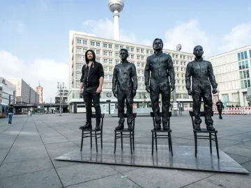 Anything To Say? sculpture in Berlin, with three bronze-like statues and a live person standing on chairs in a city plaza.
