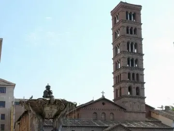 Fountain of the Tritons with sculpted figures supporting a shell-like basin, water cascading into the basin below, with a church campanile in the background.