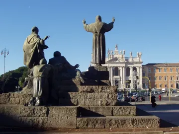 Bronze Monument to Saint Francis of Assisi on a tiered stone pedestal with a church building in the background.