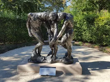 The Three Shades by Auguste Rodin, a dark bronze sculpture of three intertwined male figures with bowed heads outdoors near greenery.