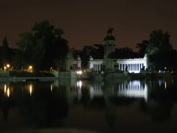 The Alfonso XII Monument lit up at night beside a calm lake, with its colonnade and equestrian statue reflected in the water.