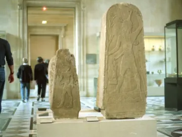 Two carved stelae on display in a museum hall, with visitors walking past in the background.