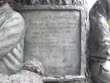 Close-up of the plaque on the Betty Campbell statue with engraved English and Welsh text, framed by sculpted children and a carved wooden trunk.