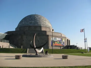 A crescent-shaped sundial sculpture with a tall gnomon stands on a paved plaza in front of a domed building.
