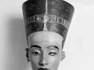 Bust of Nefertiti with a tall striped crown and decorated collar, shown in black and white.