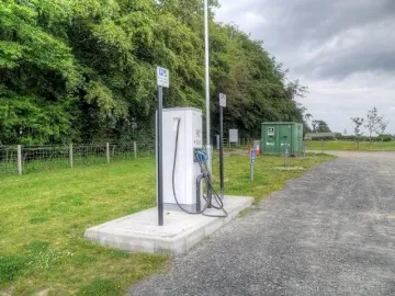 Electric vehicle charging station beside a gravel path at Northumberlandia, with trees, grass, and a utility box behind it.