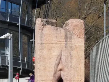 Pi-Chacán, a tall reddish-pink stone sculpture with a deep vertical cleft, standing outdoors near modern buildings and grass.