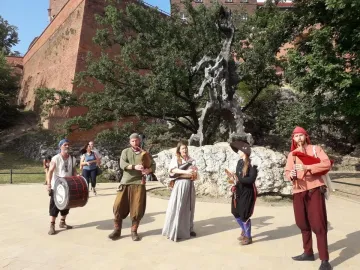 The Wawel Dragon sculpture standing on a rocky base with trees behind it and people in historical attire nearby.
