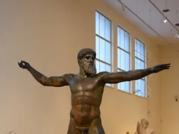 Artemision Bronze, a dark bronze statue of a nude bearded man with outstretched arms in a museum gallery.