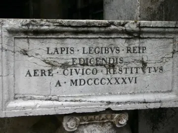 Weathered stone plaque with a carved Latin inscription and a decorative stone capital beneath it.