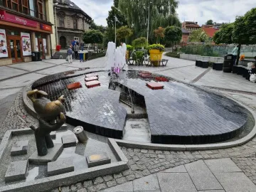 Reksio Monument beside a split egg-shaped fountain with pink-lit water in a town square.