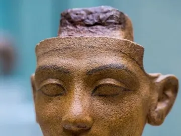 Unfinished head of Nefertiti in reddish-brown stone with a long neck and carved facial features on a light speckled base.