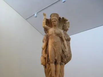 Dancers of Delphi sculpture with three draped female figures on an acanthus column in a museum gallery.