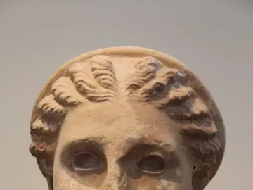Weathered stone head of Lycosoura Artemis with intricate wavy braids and a headband, mounted on a dark base.
