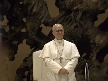 A smiling man in a white cassock stands before The Resurrection sculpture.