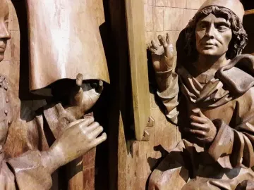 Saint Luke wooden sculpture detail showing two draped figures gesturing beside a paneled wooden background.