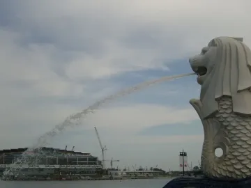 Merlion statue spraying water into the bay with a modern building and cranes in the background.