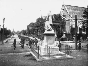 Rolleston Statue on a tall pedestal beside a fenced street, with uniformed men marching in the distance.