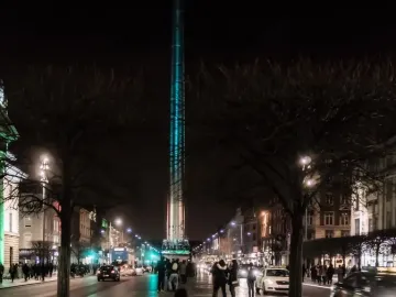 The Spire of Dublin illuminated in blue-green light at night on a city street.