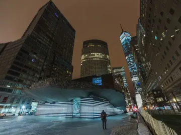 Irish Hunger Memorial with a rocky upper facade and illuminated stepped section beside downtown skyscrapers at night.