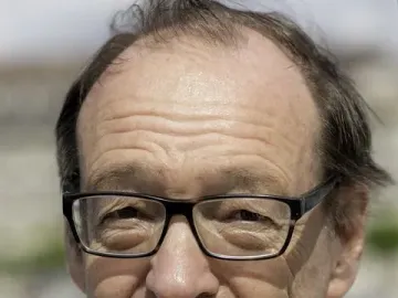 Smiling older man with black-rimmed glasses wearing a light jacket and white collared shirt.