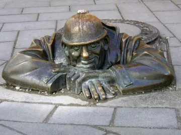 Čumil, a bronze man wearing a helmet, leans out of a manhole in a paved street.