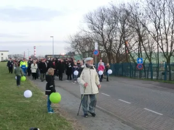 A procession of people walks along a paved path near the Kindertransport monument, carrying glowing white and green lanterns.