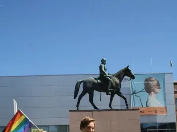 Marshal Mannerheim equestrian statue on a granite plinth with a crowd and a person in a lime green car holding a rainbow flag in the foreground.