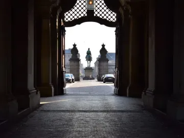 Christian IX equestrian statue seen through a dark stone archway, with a sunlit plaza and parked cars beyond.