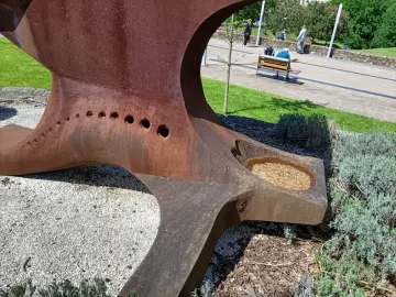 Dorna sculpture with a rusty metal surface, circular holes, and a basin filled with gravel in a park setting.