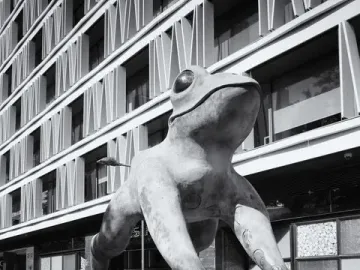 Lucky Frog sculpture on a city sidewalk beside a modern building.
