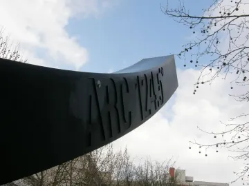 Arc de 124,5° sculpture seen from below against a partly cloudy blue sky, with bare branches framing the upper right.