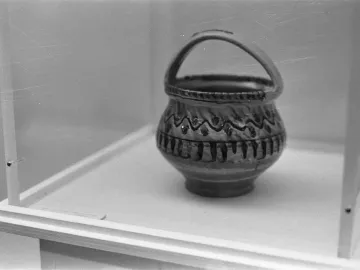 A small ornate ceramic vessel with a handle and wavy decorative patterns, displayed in a clear case.