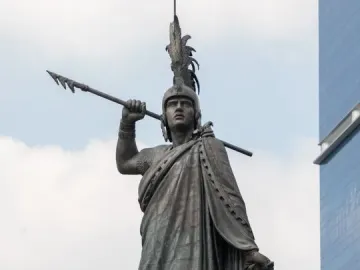 Cuauhtémoc Monument statue with a feathered headdress, spear, and document against a cloudy sky.