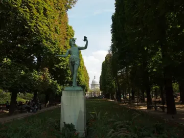The Greek Actor statue on a tall stone pedestal in a tree-lined park, with a grassy avenue and distant domed building behind it.