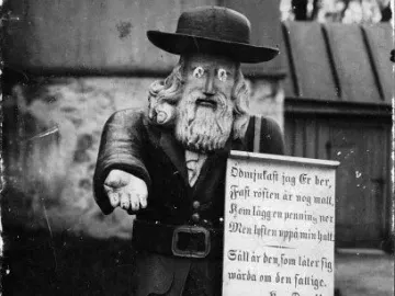 Rosenbom, a wooden bearded figure in a hat holding out one hand and a sign.