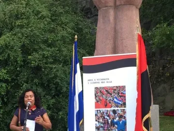 A woman in a blue dress speaks into a microphone near La Mano, with flags and a banner behind her.