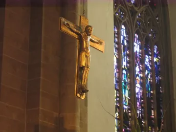 Mindener Cross hanging on a wall beside a stained-glass window in a dimly lit interior.