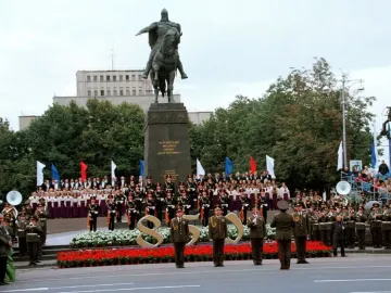 Monument to Yuri Dolgorukiy on a dark pedestal, with a crowd, choir, and band gathered in front of it.