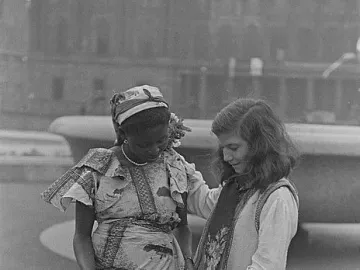 Two women examine a patterned piece of fabric or paper together, one wearing a printed dress and headscarf and the other a light outfit with a decorative vest.